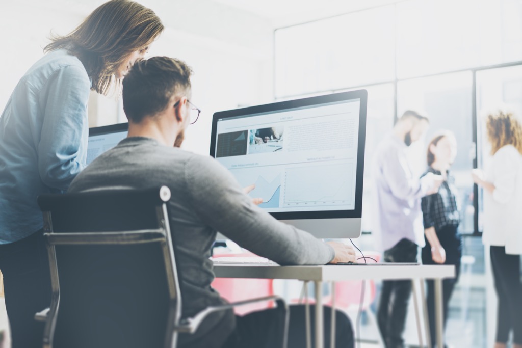 Man sitting at desk and woman standing, both of them looking at large desktop computer screen