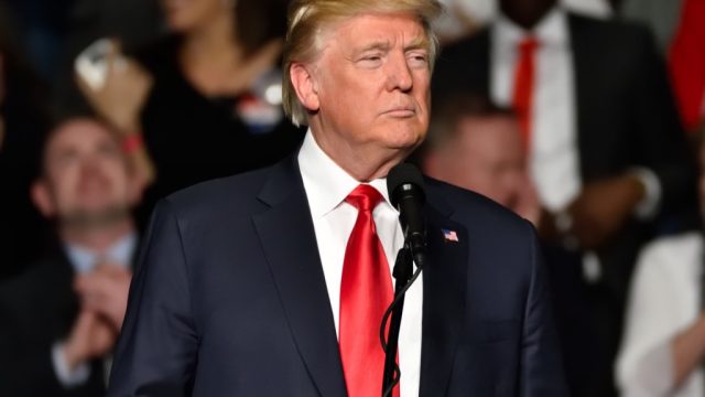 President Donald Trump stands at podium in Red tie and navy suit, set to meet prince william in 2019
