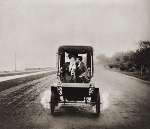 Man and woman drive down road in horseless carriage