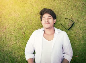 young asian man lying on the grass with his eyes closed and his sunglasses next to him