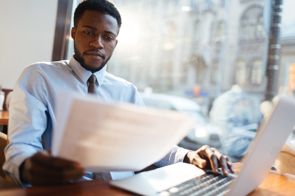 Man sitting at window in office, looking at papers and using laptop