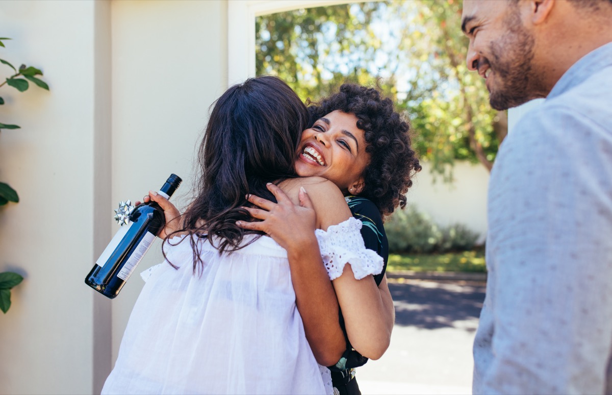 Black woman with natural hair hugs female friend at door with housewarming wine in hand, ways to feel amazing