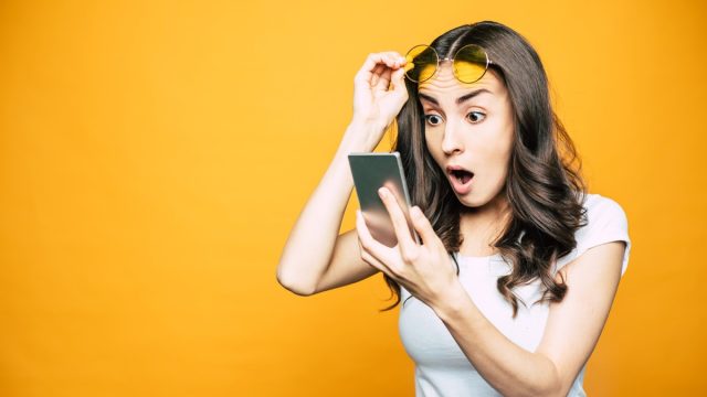 young white woman raising her sunglasses in surprise while looking at her phone