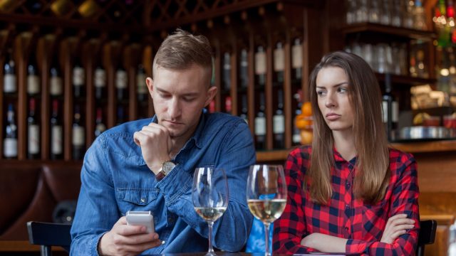young white man looking at his phone while white girlfriend looks upset at wine bar