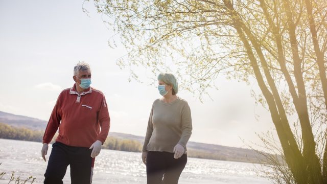 older white man and white woman walking by the water with face masks