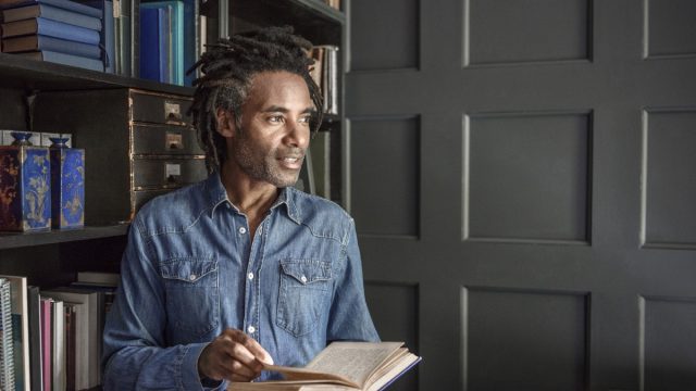 Man with dreadlocks in his 50s reading hardback in library, with bookshelves behind, pensive expression on his face