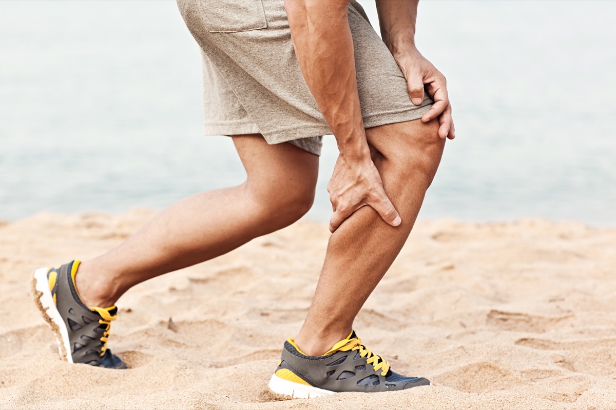 man on beach, leaning down to grab his calf with a muscle cramp