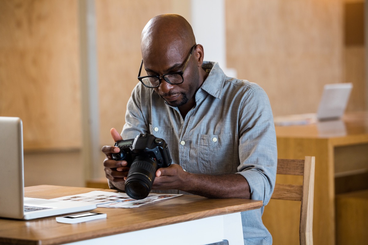 Black man in denim shirt with black glasses holds camera in front of computer police officer secrets