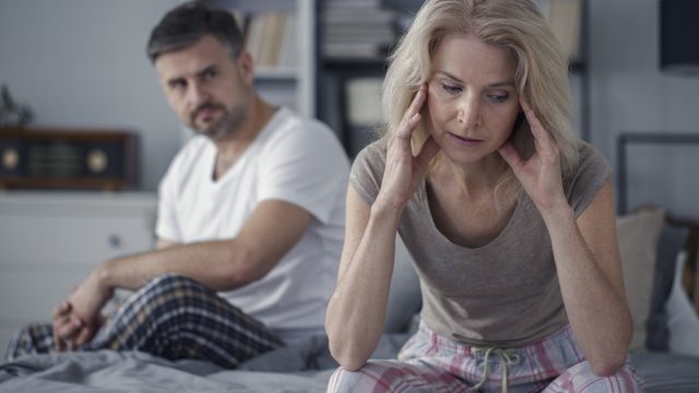 Woman holds head in hands as husband looks on in bed