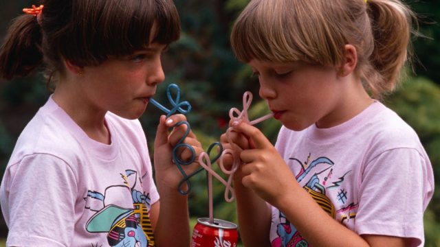 In 1980s, Two young twin sisters sip Coke from their own curly straws in an English garden, UK