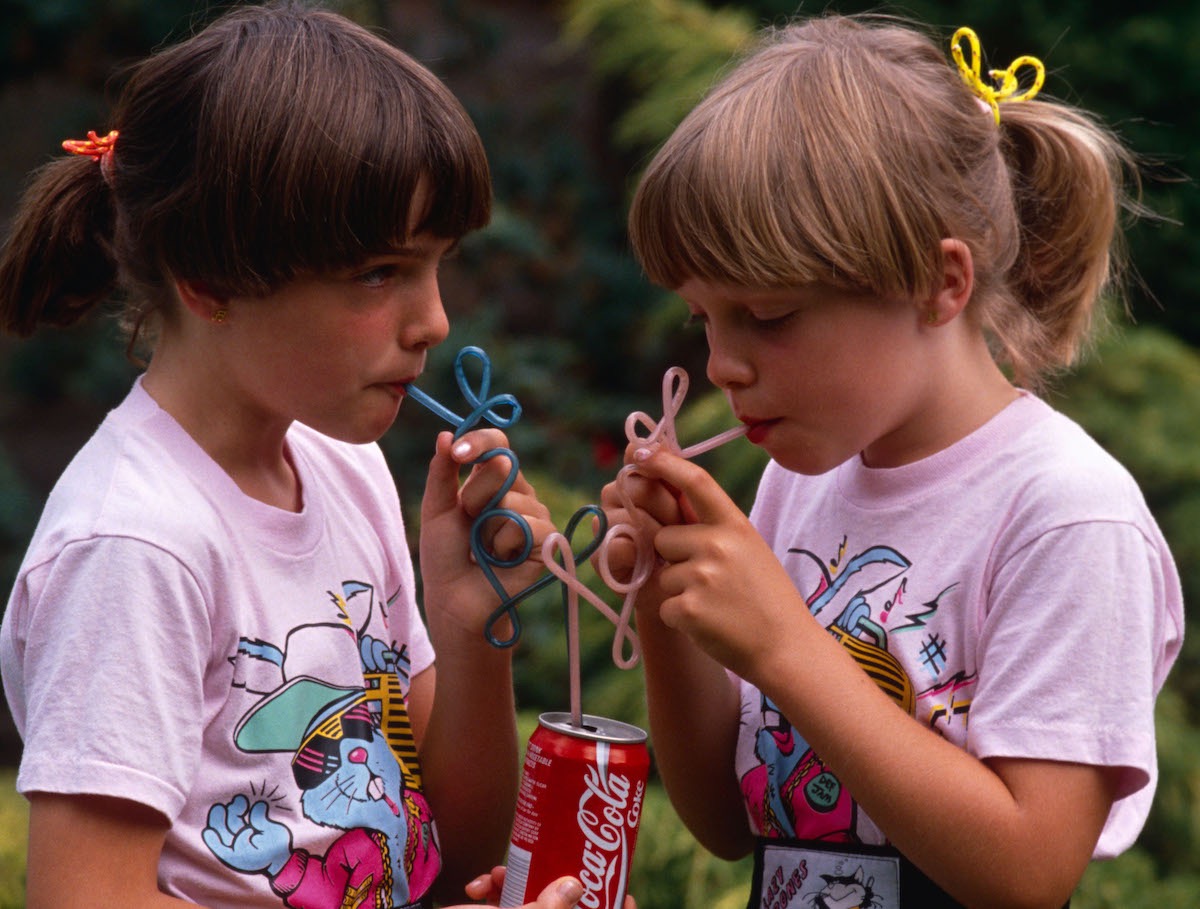 In 1980s, Two young twin sisters sip Coke from their own curly straws in an English garden, UK