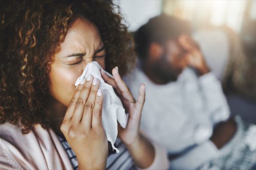Shot of a young woman blowing her nose with her boyfriend in the background