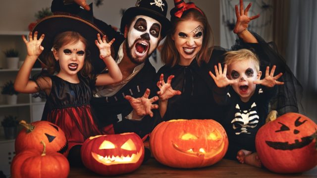 Family celebrating Halloween in traditional witch, skeleton costumes, with pumpkins in front of them