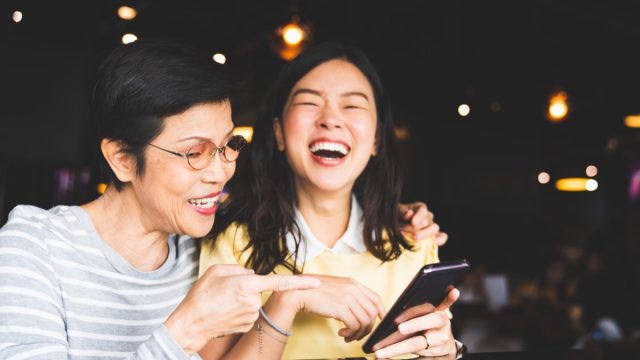 asian mother and daughter looking at a phone screen together, laughing in a cafe