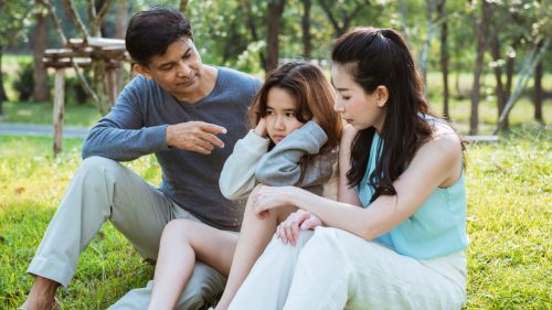 parents lecturing a female child who's covering her ears while they all sit outside