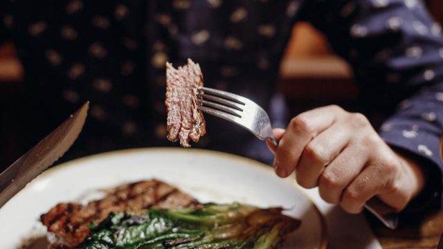 Hands holding fork and knife and eating delicious juicy steak with grilled cabbage,tomatoes and cheese on table at cafe in city street. Man tasting bbq with vegetables in restaurant