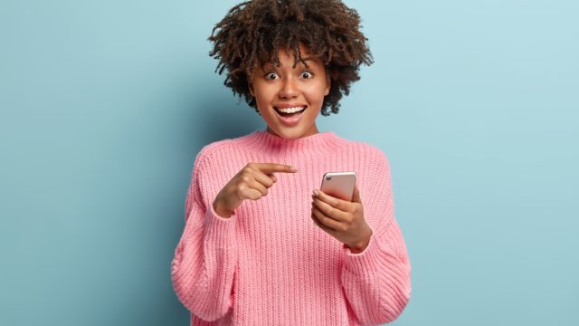 Surprised positive dark skinned lady points at smart phone device, suggests to have conversation in group chat, glad to recieve many messages of congratulation on mailbox, has overjoyed look
