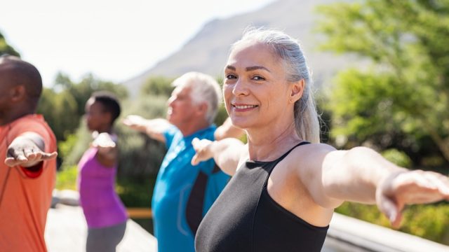 Portrait of happy senior woman practicing yoga outdoor with fitness class. Beautiful mature woman stretching her arms and looking at camera outdoor. Portrait of smiling serene lady with outstretched arms at park. (Portrait of happy senior woman pract