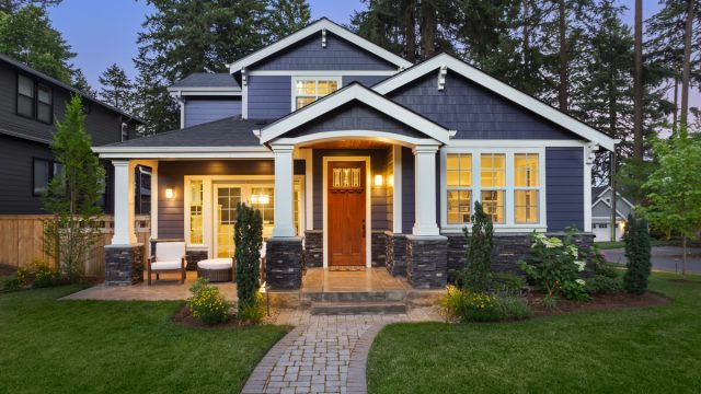facade of dark blue home with manicured lawn, landscaping, and backdrop of trees