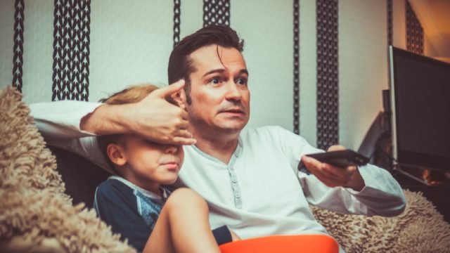 Shocked father covering son's eyes and changing channel while watching inappropriate television content at home.