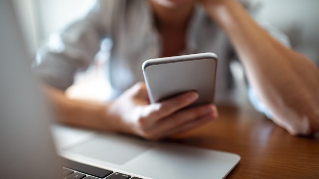 Close up of a young woman using her phone at home in front of laptop