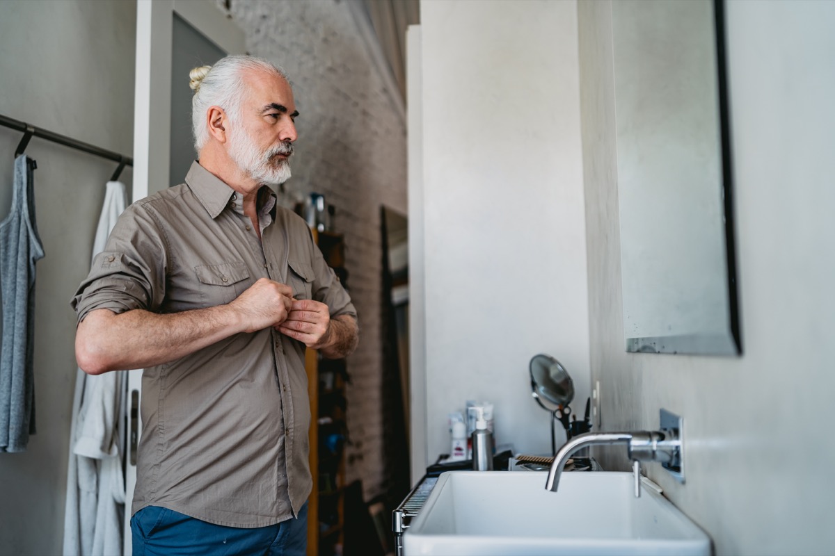 A mature man with white hair tied into a bun buttons his taupe shirt in the bathroom mirror