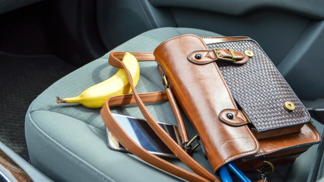 This is an image of a brown purse on the passenger seat of a car with contents spilling out. There is a pen, makeup item, phone and banana laid out around the purse. The car interior is grey.