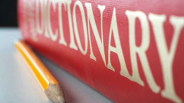 A red dictionary sitting on a table top with a pencil next to it.