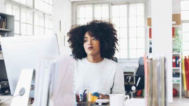 Tired young woman sitting on desk and using computer. Papers and tools, a cup on desk. Tall windows, shelves with folders, office seat on background.