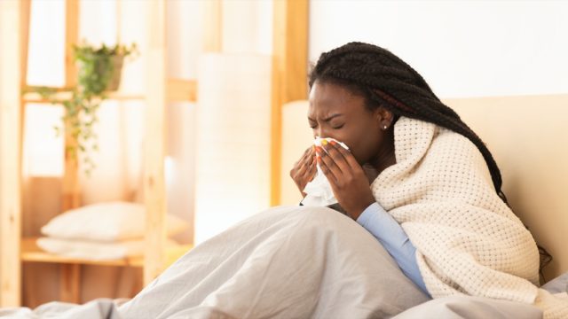 young black woman under blanket sneezing or blowing nose while sick