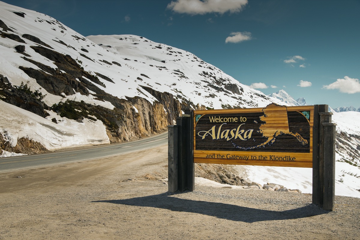 a wooden "Welcome to Alaska" in front of white mountains and off of a highway