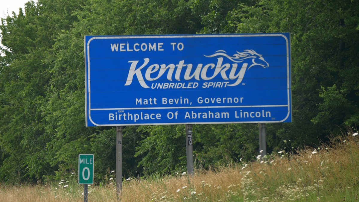 a blue "Welcome to Kentucky" sign in front of green trees