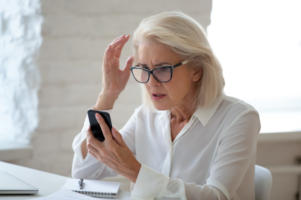 A senior woman wearing glasses and sitting at a desk looks at her smartphone with a surprised and confused look, maybe the victim of a scam.