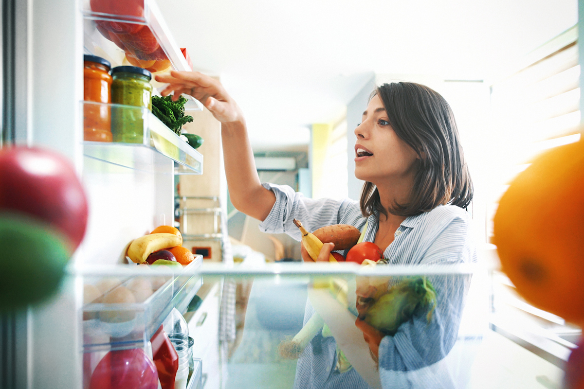 Shot from inside the fridge of a young woman picking some fruit and veggies from the fridge