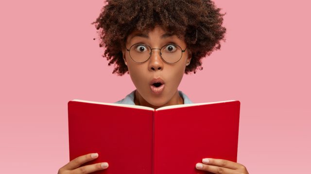 Smart young woman with glasses looking amazed while reading a book