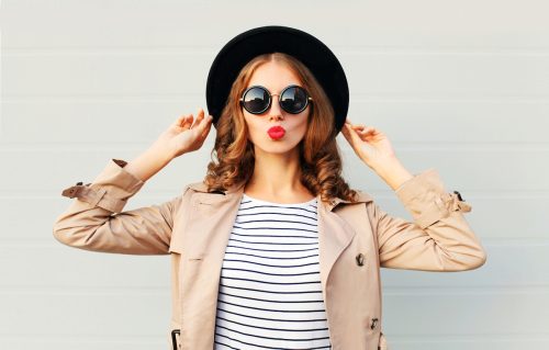 Young woman with red lipstick wearing a black hat, sunglasses, and a coat over a grey background