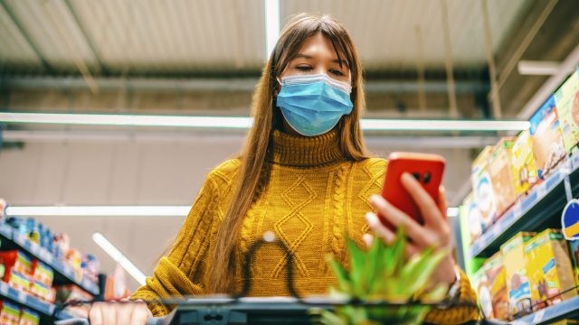 A young woman wearing a face mask looks at her phone while grocery shopping
