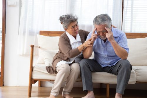 An upset older man sitting on the couch with his head in has hands while a woman comforts him