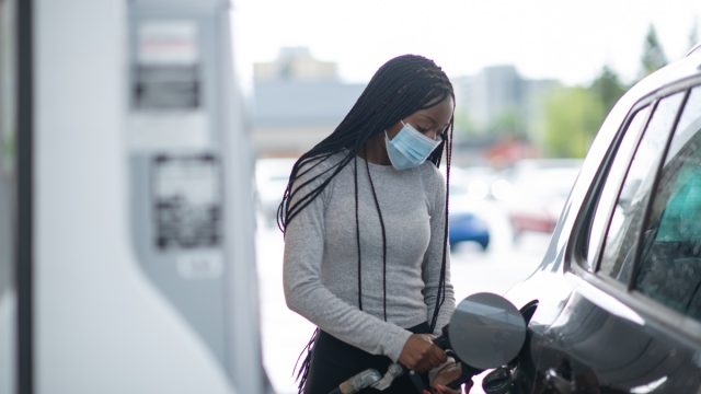 A young woman pumps gas at gas pump. She is wearing a blue medical mask and blue protective gloves.