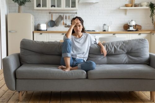 A woman sits on her couch with the kitchen in the background. She is holding her head in her hands sighing, looking exhausted
