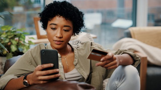 Young woman holding her credit card while using her cellphone at home