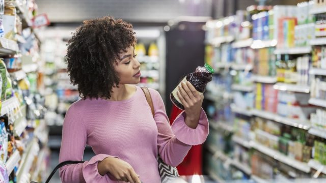 woman in her 20s shopping in a grocery store, carrying a shopping basket. She is reading the ingredient label on a bottle.