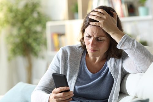 Woman with her hand on her head, looking distressed at her phone
