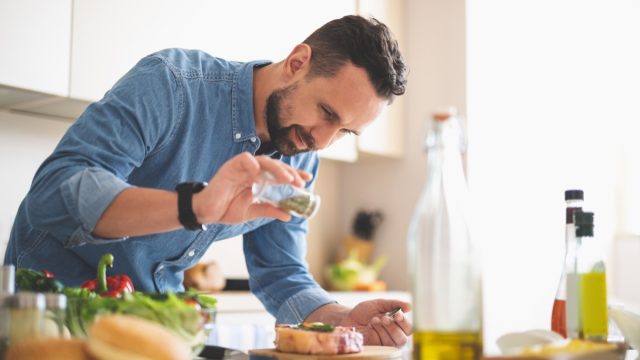 For better taste. Waist up portrait of handsome bearded gentleman in denim shirt seasoning raw beef steak