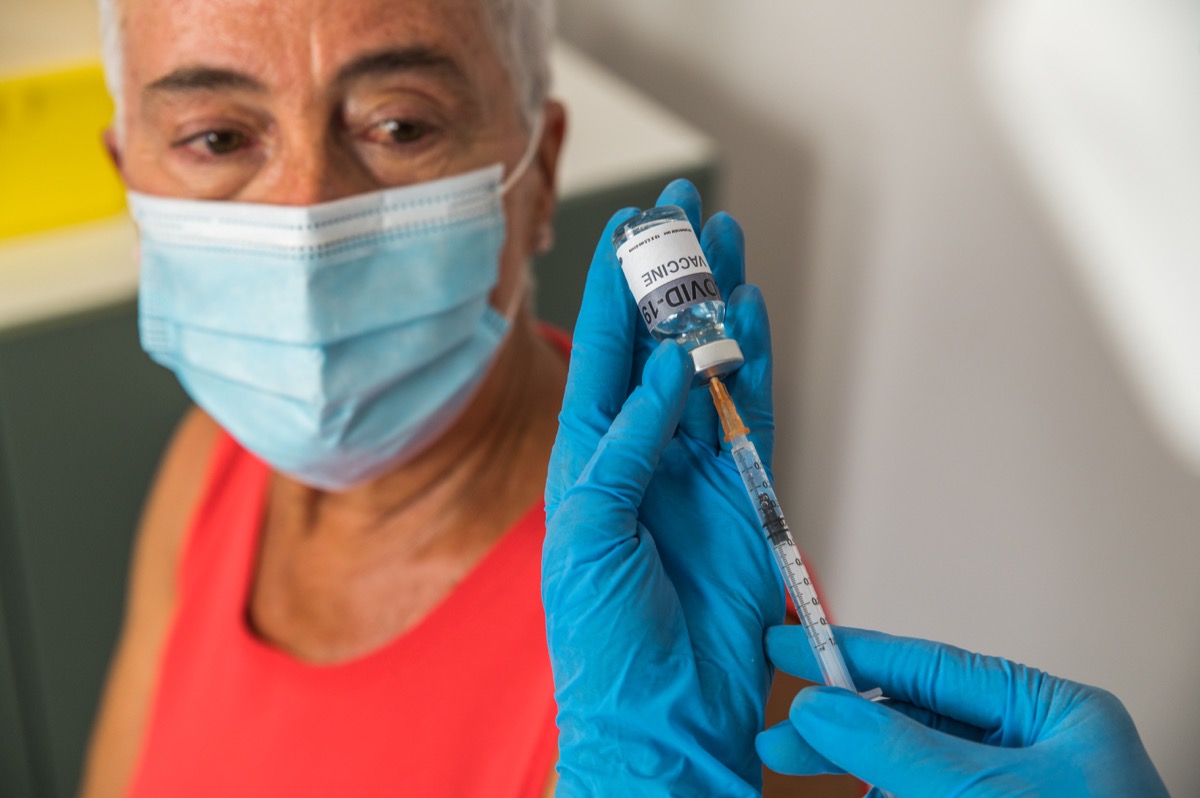 Crop anonymous doctor in gloves filling syringe with COVID 19 vaccine against blurred senior patient in hospital