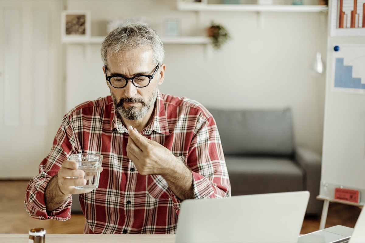Man with gray hair wearing a red plaid shirt takes a pill or supplement with a glass of water