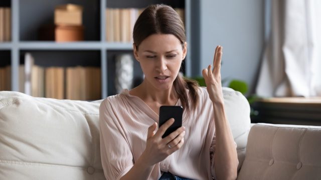 Confused angry woman having problem with phone, sitting on couch at home, unhappy young female looking at screen, dissatisfied by discharged or broken smartphone, reading bad news in message