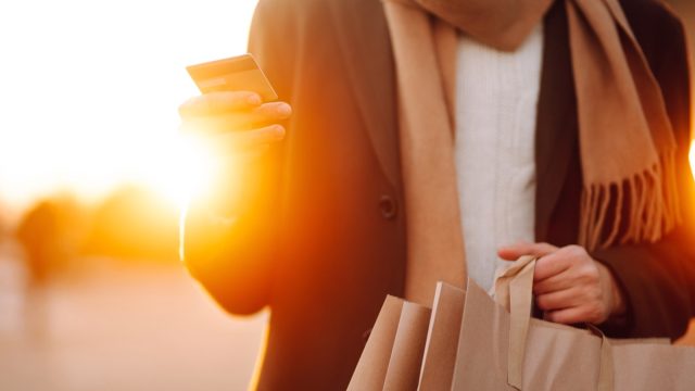 Smiling man with shopping bags enjoying in shopping at sunset. Young man with phone walking down the street. Purchases, black friday, discounts, sale concept. Autumn shopping.