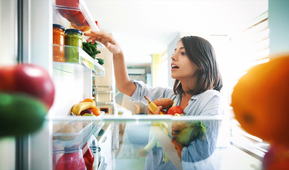 Young woman with her hands full of fruits and veggies, organizing her fridge