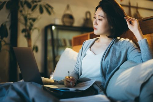 woman sitting on the bed, working from home using laptop and handling paperworks
